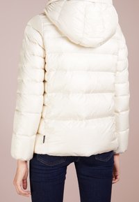 Vit puffjacka med huva, quiltat mönster och elastiska ärmslut. Slät textur med en figurnära siluett. Matchad med mörkblå jeans.