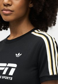 Κοντινό πλάνο ατόμου που φοράει μαύρο αθλητικό πουκάμισο Adidas με λευκές και κίτρινες ρίγες στα μανίκια και λογότυπο στο στήθος.