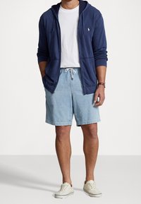 Marin zip-up hoodie, vit t-shirt, ljusblå denimshorts och beiga sneakers. Hoodien har en liten logotyp på bröstet; avslappnad passform.