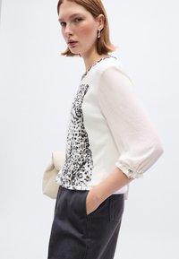 Λευκό πουκάμισο με μοτίβο μαύρου λεοπάρ, μανίκια τριών τετάρτων με σούρες στις μανσέτες, συνδυασμένο με σκούρο γκρι παντελόνι.