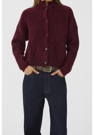 Kvinde iført en knappet bordeauxfarvet fluffy cardigan, mørkeblå højtaljede jeans og et brunt bælte med hænderne i lommerne.