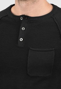 Zwarte gestructureerde henley shirt met ronde hals, een knoopsluiting met drie knopen en een klein zakje op de linkerbovenkant. Beschikt over ribgebreide stof.