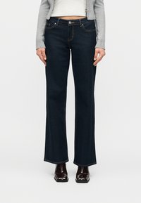 Γυναίκα που φοράει σκούρα μπλε τζιν με ψηλή μέση και καμπάνα στα μπατζάκια, ένα ανοιχτό γκρι κοντοκουμπωτό ζακέτα και σκούρα καφέ μπότες με τετράγωνη μύτη.