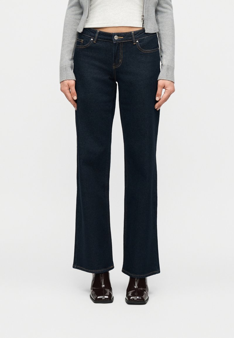 Γυναίκα που φοράει σκούρα μπλε τζιν με ψηλή μέση και καμπάνα στα μπατζάκια, ένα ανοιχτό γκρι κοντοκουμπωτό ζακέτα και σκούρα καφέ μπότες με τετράγωνη μύτη.