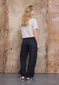 Kvinde med langt blondt hår står med front mod en trævæg iført en lysegrå t-shirt med korte ærmer, sorte bukser og brune sneakers.