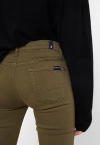 Jeans de color verde oliva con un diseño ajustado, que cuentan con un parche de tela más oscura en el bolsillo trasero y un sutil detalle de costura.