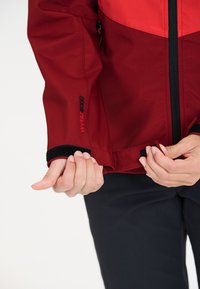 Red and dark red jacket with a zip front, velcro cuffs, and "Waterproof 5000" printed on the sleeve. Smooth fabric with a fitted design.