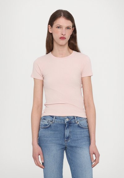 Mulher com longos cabelos castanhos, vestindo uma camisola curta de malha de cor rosa claro e calças de ganga azuis, em pé contra um fundo branco simples.