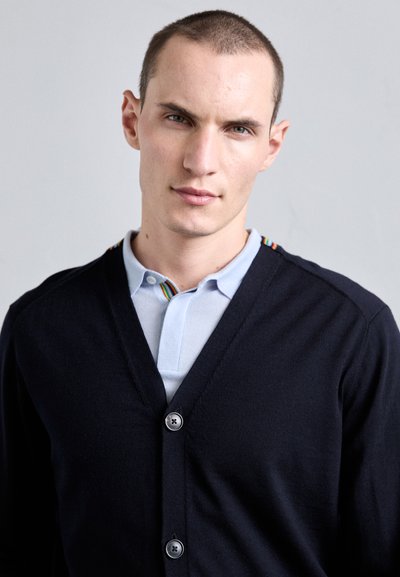 Marineblå cardigan med V-hals og fem knapper, båret over en lyseblå polo. Stripet detalj på kragen tilføjer karakter til outfittet.