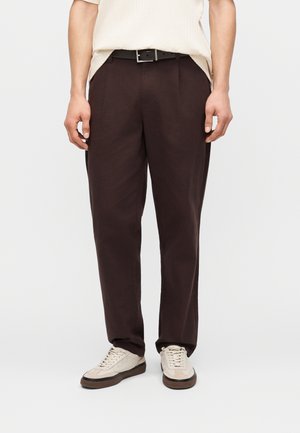 Mężczyzna ubrany w ciemnobrązowe spodnie, czarny pasek, kremową koszulę z krótkim rękawem oraz beżowe tenisówki, stojący na białym tle.