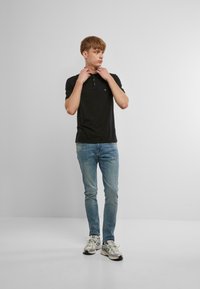 Fekete póló ing, gombos kivágással, világoskék farmernadrággal párosítva. A modell szürke és fehér sportcipőt visel, semleges háttér előtt áll.