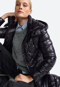 Sort, skinnende pufferjakke med hætte, der har et quiltet design og lynlås. Bæres over en grå sweater og en blåstribet skjorte med krave.