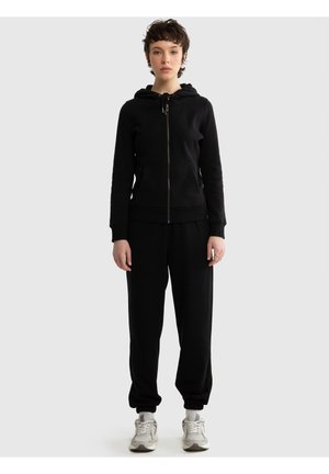 Noor inimene seisab, kandes musta tõmblukuga kapuutsiga pusa, musti pükse ja valgeid tossusid heleda halli tausta ees.