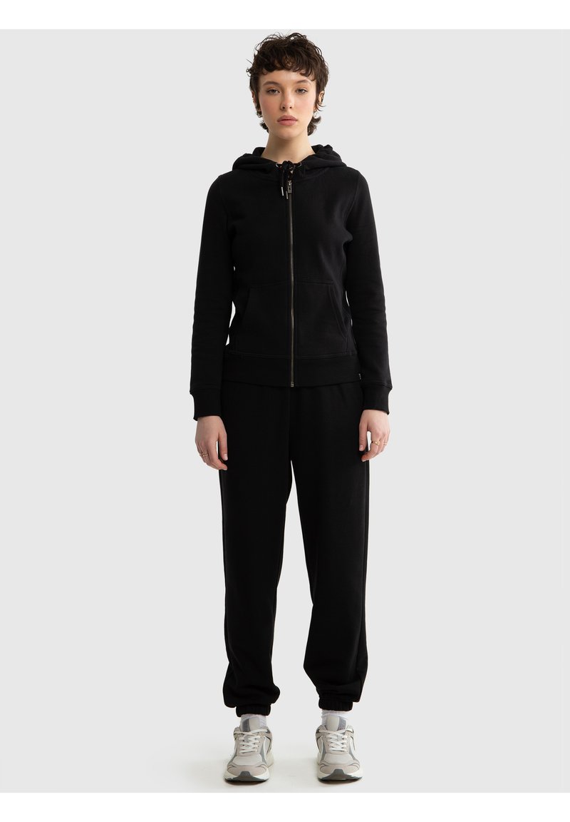 Tânăr în picioare, purtând un hanorac negru cu fermoar, pantaloni sport negri și adidași albi, pe un fundal simplu, gri deschis.