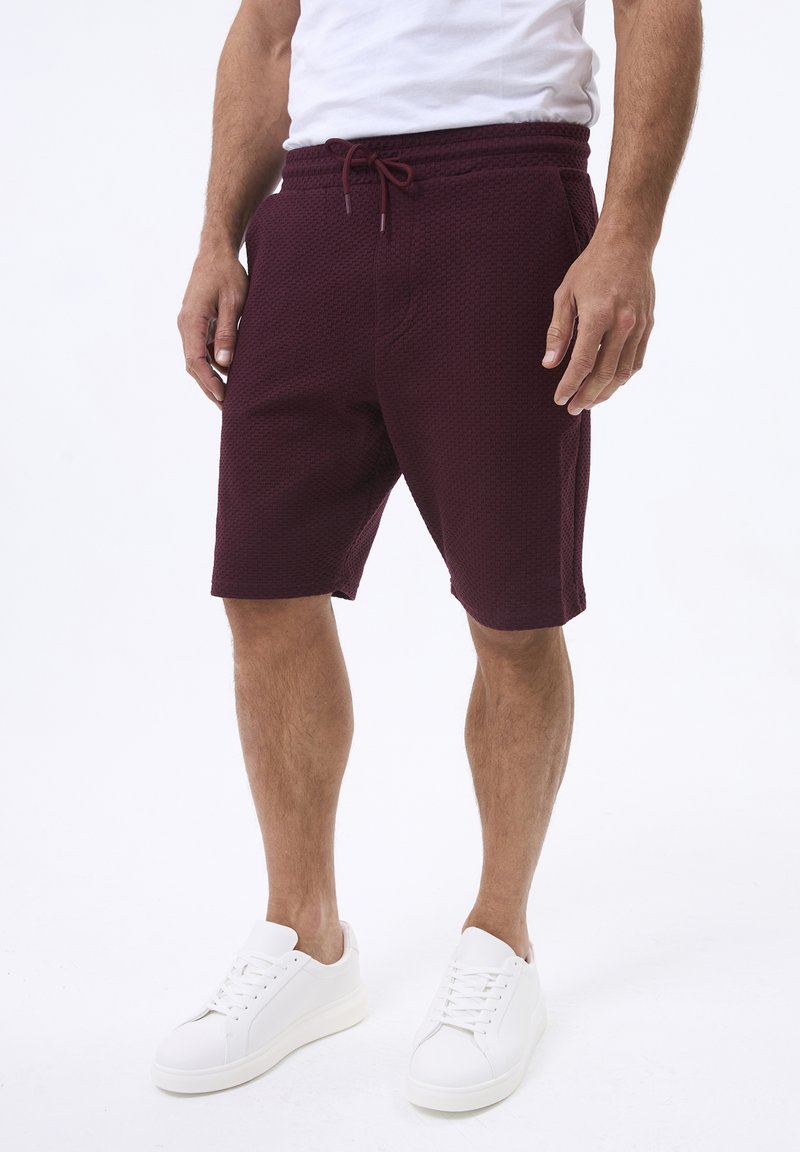 Shorts bordeaux texture con vita elastica e cordoncino, dotati di tasche laterali, abbinati a sneakers bianche.