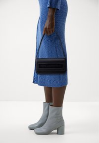 Zwarte leren handtas met een reliëflogo, rechthoekige vorm en een schouderriem. Draagt bij een blauwe ribgebreide jurk en lichtgrijze enkellaarzen.