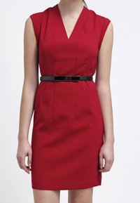 Červené šaty bez rukávů s výstřihem do V, přiléhavý střih a černý pásek v pase. Hladká látka s minimálním švem a rovný střih.