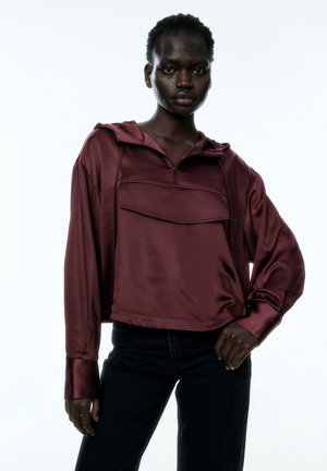 Bordeaux satijnen hoodie met een kwart rits, voorzien van een voorzak en lange mouwen, gecombineerd met donkere jeans.