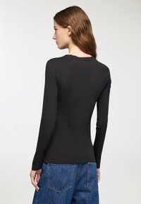 Donna con lunghi capelli castani che indossa una maglietta nera attillata a maniche lunghe e jeans blu, in piedi e di spalle contro uno sfondo chiaro.