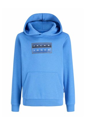 Adolescent aux cheveux bouclés portant un sweat à capuche Jack & Jones bleu clair, un jean noir et des baskets noires, debout et souriant sur un fond uni.