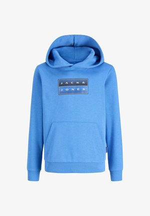 Adolescent aux cheveux bouclés portant un sweat à capuche Jack & Jones bleu clair, un jean noir et des baskets noires, debout et souriant sur un fond uni.