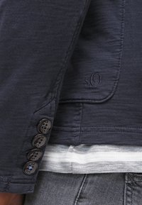 Primer plano de la manga de una chaqueta azul marino con cuatro botones y el logo bordado "s.O" encima de una capa de tela a rayas y pantalones grises.