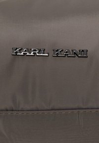 Tamsiai pilka striukė, pasižyminti lygiu, matinio paviršiaus audiniu, su išsiskiriančiu juodu metaliniu logotipu "KARL KANI" ant krūtinės.