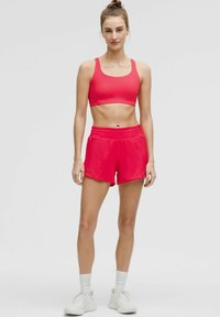 Sostenedor deportivo rojo y pantalones cortos a juego, hechos de material elástico. Los pantalones cortos tienen una banda elástica en la cintura, y el modelo lleva zapatillas blancas con calcetines.