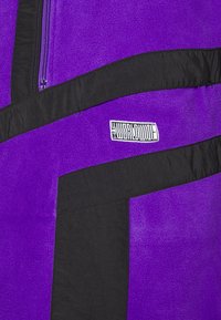 Lilla fleecejakke med sorte detaljer, der har en lynlås lomme, abstrakte designmønstre og et broderet logo-lap.