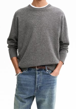 Homme portant un pull gris à col rond associé à un jean bleu et une ceinture marron, les mains dans les poches avant.