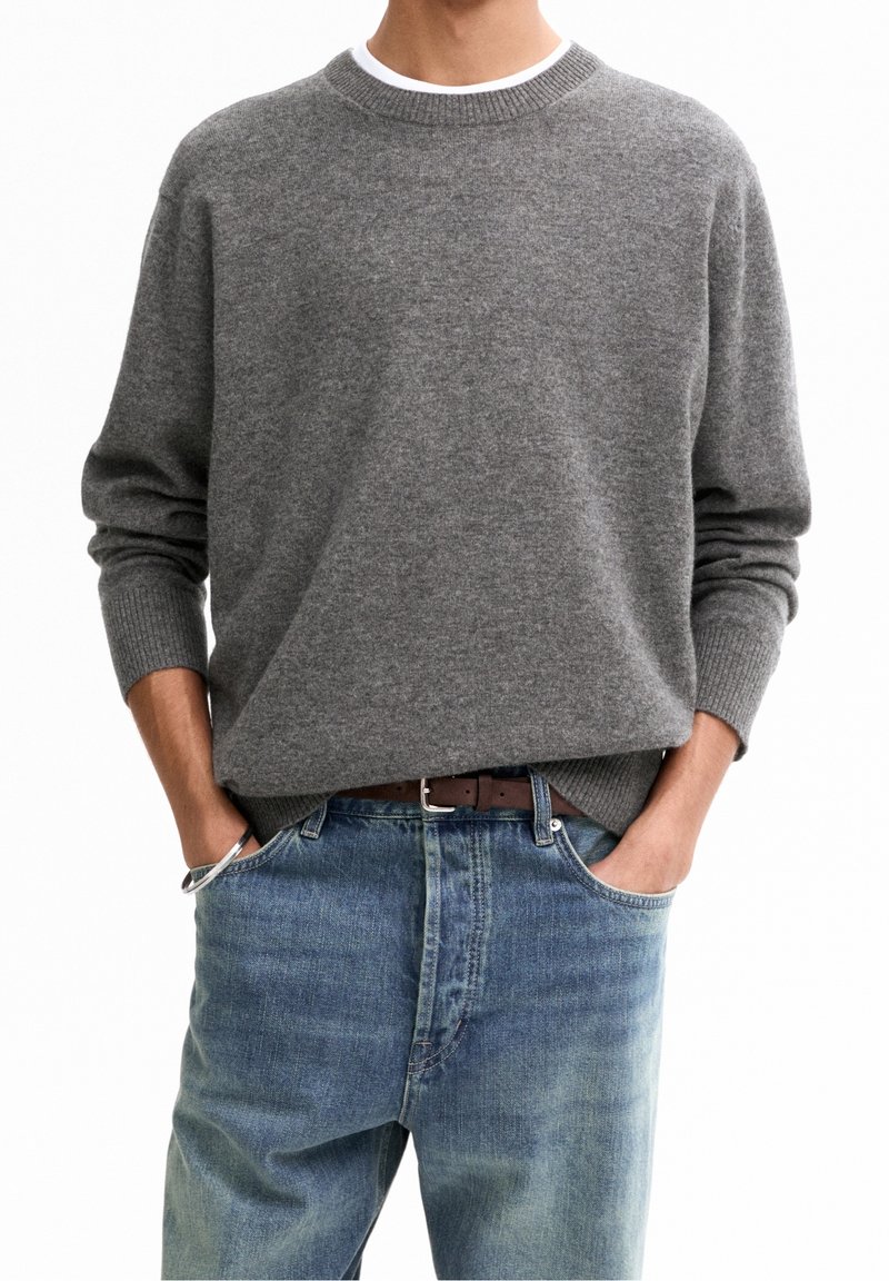 Homme portant un pull gris à col rond associé à un jean bleu et une ceinture marron, les mains dans les poches avant.