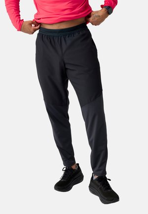 Oseba, oblečena v črne športne hlače in črne superge, si prilagaja pas, medtem ko stoji ob preprostem ozadju.