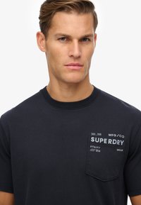 Nahaufnahme eines Mannes, der ein schwarzes Superdry-T-Shirt mit Brusttasche und weißem, aufgedrucktem Text auf der Tasche vor weißem Hintergrund trägt.