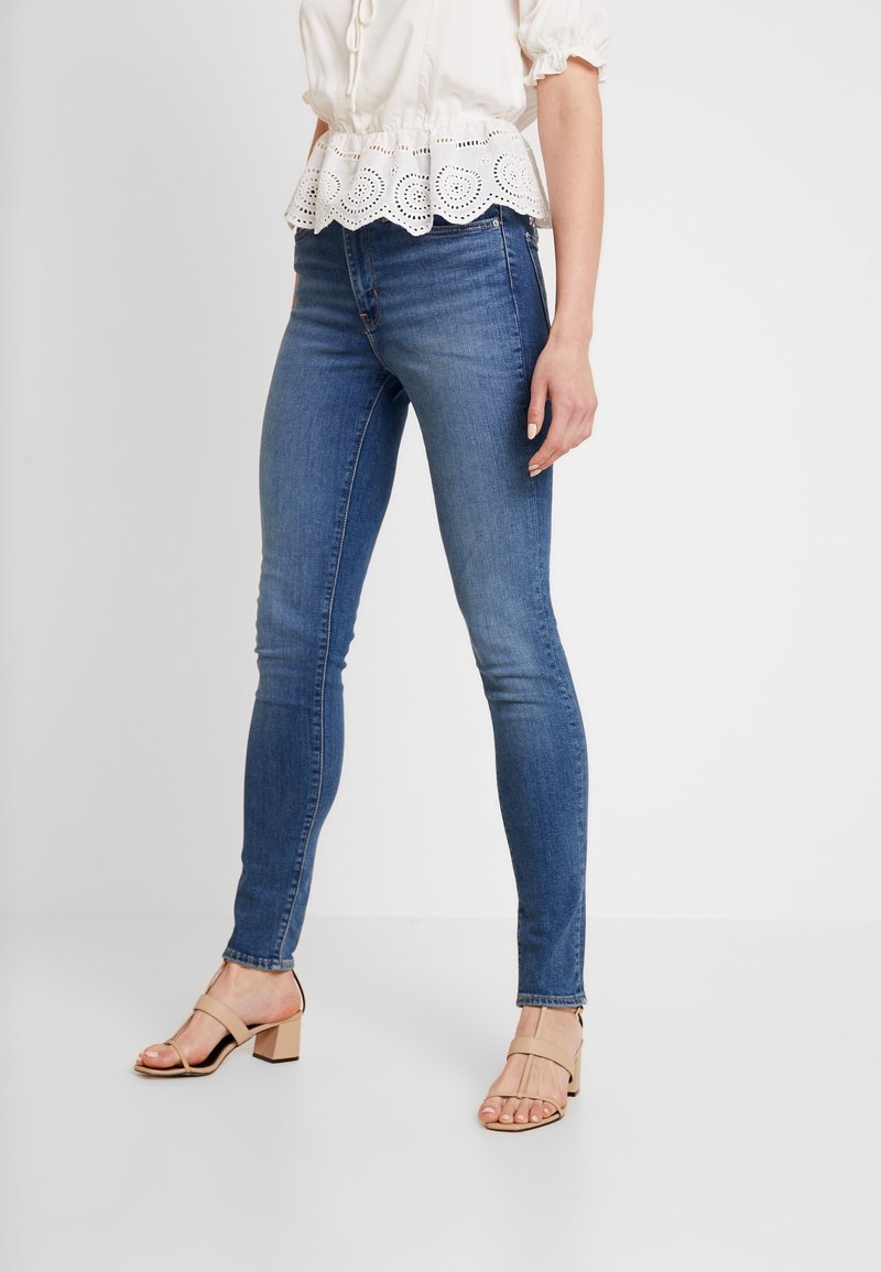Jeans skinny en denim bleu moyen, avec une texture lisse et une silhouette ajustée, associés à un chemisier blanc avec une bordure brodée ajourée.