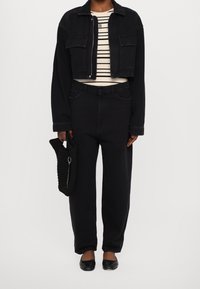 Chaqueta de mezclilla negra corta sobre una camisa de rayas de color claro. Jeans negros sueltos y zapatos planos negros. Bolsa tejida negra con un acento de llavero.