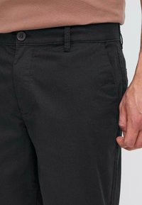 Black cotton trousers featuring a flat front, side pockets, and a single button closure. Smooth texture with minimal stitching details.