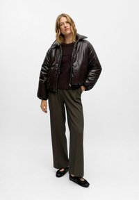 Braune, glänzende Pufferjacke mit einem breiten Kragen, getragen über einem dunklen Strickpullover und kombiniert mit hochtaillierten, geraden Hosen. Schwarze Schuhe.