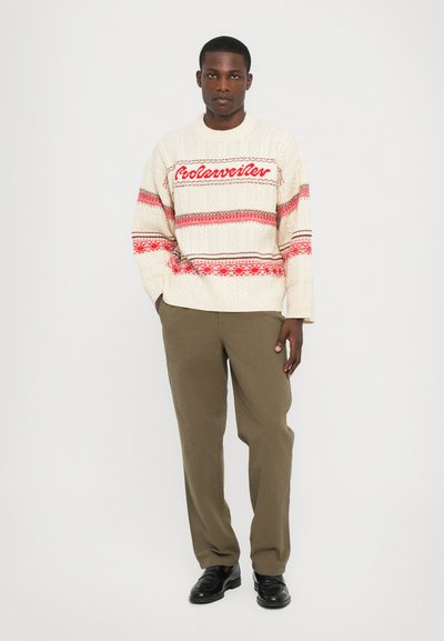 Maglione lavorato a maglia crema con accenti rossi e decorazioni, caratterizzato da un design a treccia. Abbinato a pantaloni verde oliva e scarpe nere.