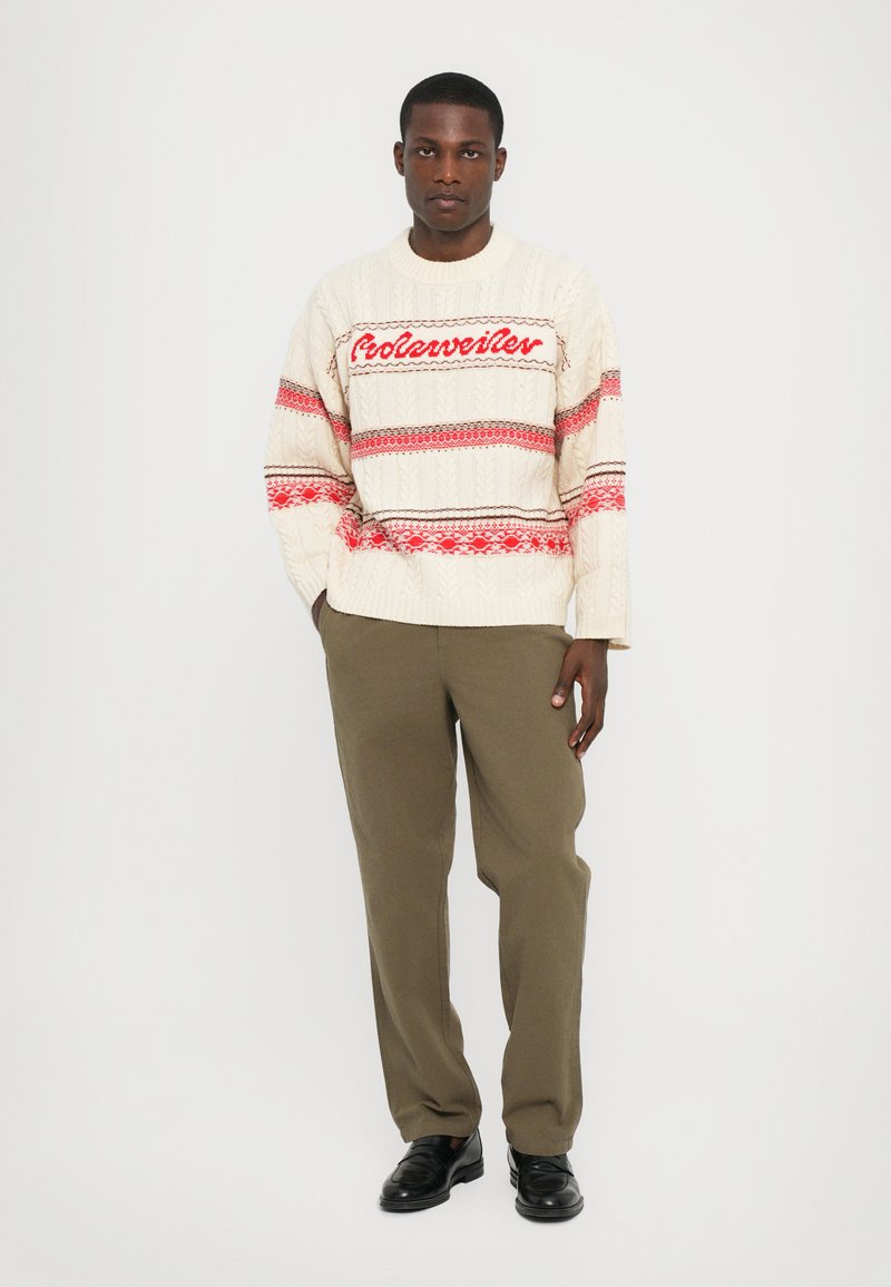 Suéter de punto crema con acentos rojos y de patrón, que presenta un diseño de punto de cable. Combinado con pantalones verde oliva y zapatos negros.