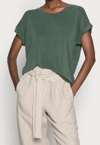 Grön kortärmad t-shirt med rund halsringning och uppvikta ärmar, tillsammans med beiga linnebyxor med hög midja, knytbälte och veck.