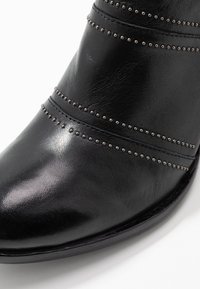 Botín de cuero negro que presenta tachuelas elevadas a lo largo de las costuras horizontales, un acabado liso y una puntera redondeada.