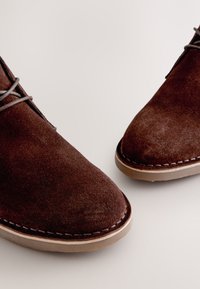 Braune Wildleder-Stiefeletten mit rund zulaufender Spitze, grauen Schnürsenkeln und einer hellbraun abgenähten Gummisohle. Nahaufnahme betont die Struktur.