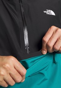 Giacca leggera nera con zip parziale frontale e logo, abbinata a una fascia in teal. Texture liscia e vestibilità atletica sono visibili.