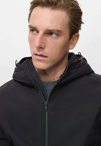 Crna zip-up jakna s kapuljačom, s glatkom teksturom i suptilnim detaljima. Nosi se preko sive vune, okrenuta malo sa strane.