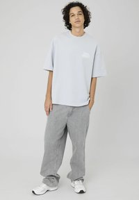 T-shirt oversize gris clair avec des manches courtes et un logo sur la poitrine gauche, associé à un jean gris ample et des chaussures de sport blanches.