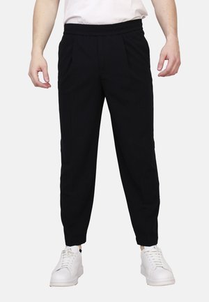 Persona che indossa pantaloni neri con taglio a sigaretta, sneakers bianche e una camicia bianca, in piedi con le braccia rilassate ai lati davanti a uno sfondo semplice.