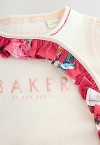 Kremowy top z falbanką w kwiatowy wzór wzdłuż dekoltu. Napis "BAKER BY TED BAKER" w miękkim różu w przodzie. Miękka bawełniana tekstura.