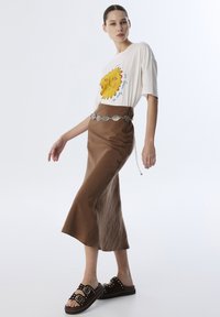 Camiseta blanca con un gráfico de un sol amarillo, combinada con una falda de satén marrón y sandalias negras tipo slide con tachuelas metálicas y detalles de doble hebilla.