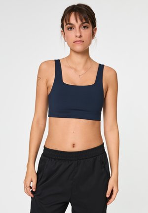 Žena v tmavomodrom športovom podprsenke a čiernych elastických športových nohaviciach, stojaca pred jednoliatym bielym pozadím.