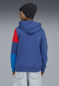La felpa con cappuccio presenta una combinazione di sezioni blu, rosse e azzurre, un tessuto testurizzato e polsini a costine. Branding minimale sul cappuccio.